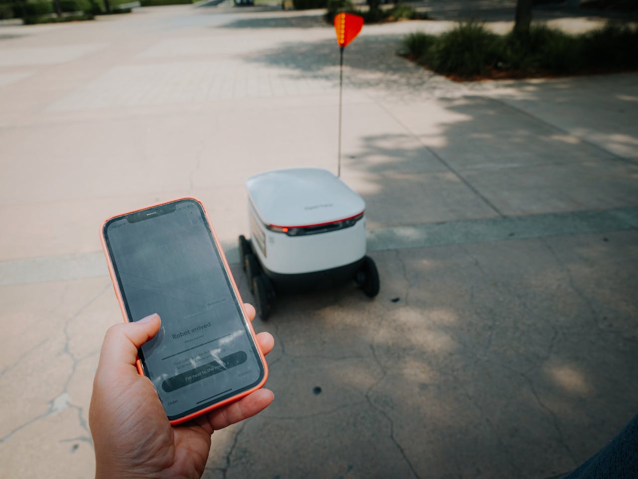 A person uses a smartphone to track an autonomous delivery robot in an outdoor setting.